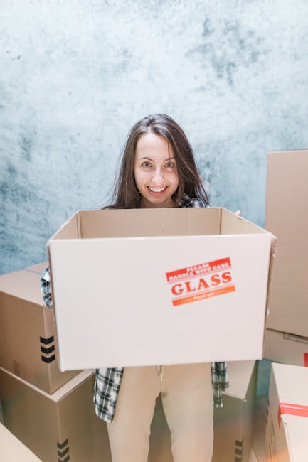 A young woman with long brown hair, smiling, standing indoors against a textured grey wall. She is holding an empty cardboard moving box labeled 'FRAGILE GLASS,' with other boxes stacked nearby on the floor. The boxes are made of corrugated cardboard, some wrapped with plastic or tape, and are arranged around her in a space that appears to be preparing for a home relocation or moving process. The scene highlights the packing stage of a house move, with natural lighting illuminating the area. The setting suggests an organized packing and loading process managed by Movers Edgware, specializing in house removals and furniture transport, supporting efficient and professional moving services.