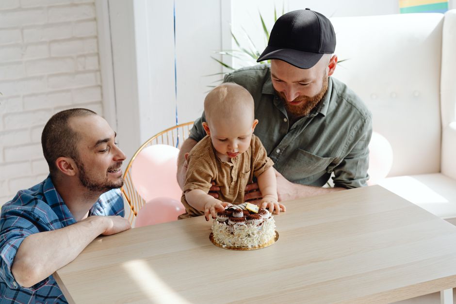 A young child with a bald head wearing a light brown shirt is sitting at a wooden table, reaching out to touch a small, round birthday cake decorated with white icing, chocolate pieces, and a yellow candle. An adult man with a beard, wearing a grey shirt and a black cap, is standing behind the child, leaning over and supporting the child’s arm. Another man, also with a beard, dressed in a blue plaid shirt, is seated at the table opposite the child, smiling and watching the scene. The setting appears to be inside a well-lit room with a white brick wall, a white sofa in the background, and pastel pink balloons tied to a chair, indicating a celebratory occasion. The image captures a moment of home celebration, possibly during a family move or house warming, with the focus on familiar surroundings. Movers Edgware, a company specialising in removals, is related to the context of this scene, which involves packing, moving, or settling into a new home environment.