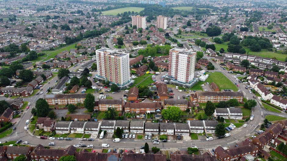 Photograph of a three-storey residential building with multiple balconies, located in a residential area under a bright blue sky with scattered white clouds. The building has a brick exterior with whi