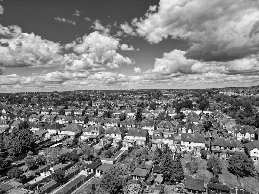 Black and white aerial view of a residential area showing numerous closely spaced houses with pitched roofs, gardens, and backyards. The scene includes outdoor furniture, fences, and pathways between properties, with some houses featuring small sheds or outbuildings. The sky above is partly cloudy with large, fluffy clouds casting shadows across the neighborhood. In the background, distant buildings and trees are visible on the horizon. This image represents a typical suburban setting suitable for home relocation activities, with the residential environment ready for household packing, furniture transport, and moving logistics. The aerial perspective emphasizes the scale and layout of the houses, as might be seen during a house removal or moving services operation carried out by professionals such as Movers Edgware, serving local areas like Edgware High Street and HA8.