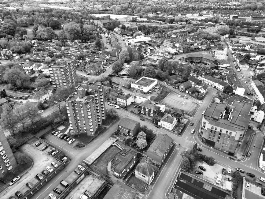 A black and white aerial photograph showing a residential and commercial area in Edgware, including multiple multi-storey apartment buildings with balconies, smaller houses, and a mix of retail and office structures. In the foreground, a paved parking lot contains numerous parked cars alongside a low-rise building with a flat roof. Beyond the parking area, streets lined with trees and vehicles lead to various properties, with some buildings featuring pitched roofs and others flat roofs. The scene captures the urban layout of Edgware, with a mix of green spaces and dense development, illustrating the typical environment for house removals and furniture transport services by Movers Edgware, who operate in the HA8 region. The lighting suggests daytime with natural light casting minimal shadows, emphasizing clear visibility of the buildings, vehicles, and surrounding infrastructure involved in home relocation and moving logistics.