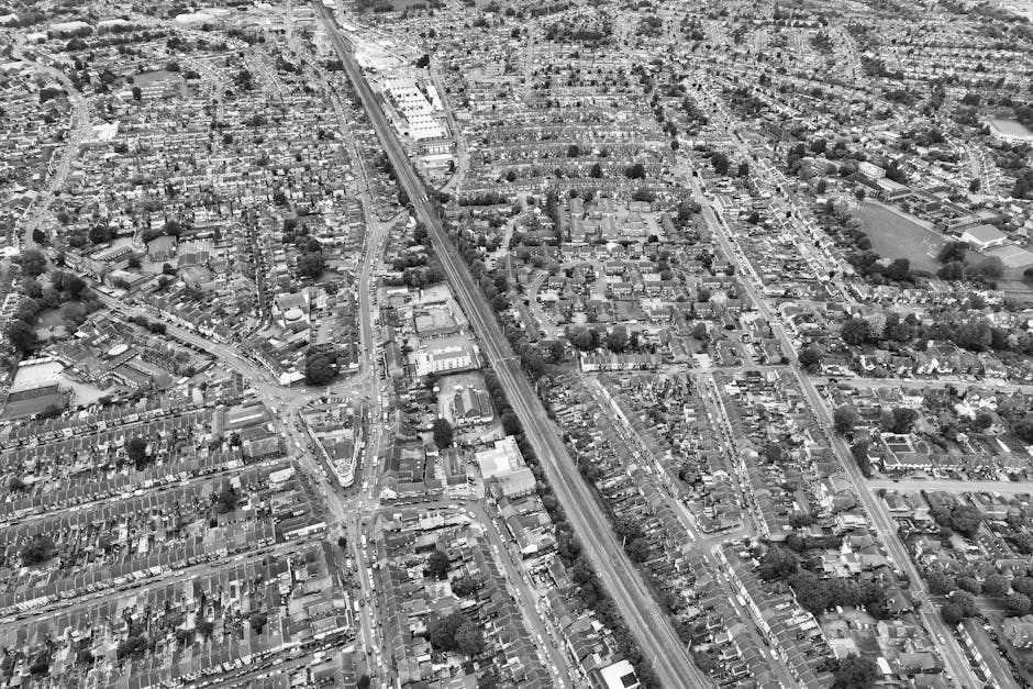 Black and white aerial view of a residential area showing numerous closely spaced houses with pitched roofs, gardens, and backyards. The scene includes outdoor furniture, fences, and pathways between properties, with some houses featuring small sheds or outbuildings. The sky above is partly cloudy with large, fluffy clouds casting shadows across the neighborhood. In the background, distant buildings and trees are visible on the horizon. This image represents a typical suburban setting suitable for home relocation activities, with the residential environment ready for household packing, furniture transport, and moving logistics. The aerial perspective emphasizes the scale and layout of the houses, as might be seen during a house removal or moving services operation carried out by professionals such as Movers Edgware, serving local areas like Edgware High Street and HA8.