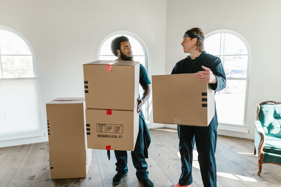Two professional movers from Movers Edgware are inside a bright, spacious room with large arched windows allowing natural light to fill the space. They are engaged in the home relocation process, each carrying a large cardboard box, which appears to be part of packing and furniture transport. The mover on the left, with curly hair and a beard, is holding a box vertically, while the other, wearing a headband and with lighter hair, is carrying a box at chest height. The room has a wooden floor and minimal furniture, including a vintage-style armchair with green upholstery positioned to the right, and a few more cardboard boxes are stacked on the floor nearby, awaiting loading. The scene captures a step in the loading process, with the movers preparing items for transport, emphasizing careful handling and organized packing. This image illustrates the stage of furniture transport and logistics involved in a professional house removal, as carried out by Movers Edgware, on their website page discussing hidden moving charges in Edgware.
