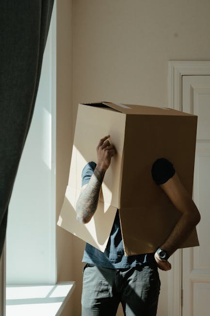 Two professional movers from Movers Edgware are inside a bright, spacious room with large arched windows allowing natural light to fill the space. They are engaged in the home relocation process, each carrying a large cardboard box, which appears to be part of packing and furniture transport. The mover on the left, with curly hair and a beard, is holding a box vertically, while the other, wearing a headband and with lighter hair, is carrying a box at chest height. The room has a wooden floor and minimal furniture, including a vintage-style armchair with green upholstery positioned to the right, and a few more cardboard boxes are stacked on the floor nearby, awaiting loading. The scene captures a step in the loading process, with the movers preparing items for transport, emphasizing careful handling and organized packing. This image illustrates the stage of furniture transport and logistics involved in a professional house removal, as carried out by Movers Edgware, on their website page discussing hidden moving charges in Edgware.